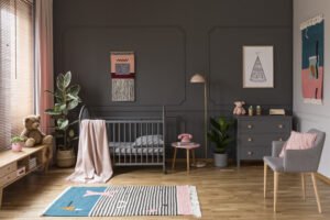Real photo of a grey crib standing next to a pink stool, a lamp and cupboard in grey baby room interior also with armchair, rug and posters